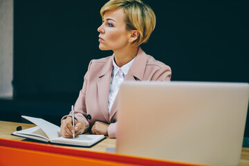 Wall Mural - Thoughtful female entrepreneur 50 years old thinking on ideas for creating successful business plan working at laptop device in office.Pondering mature businesswoman making notes in notebook