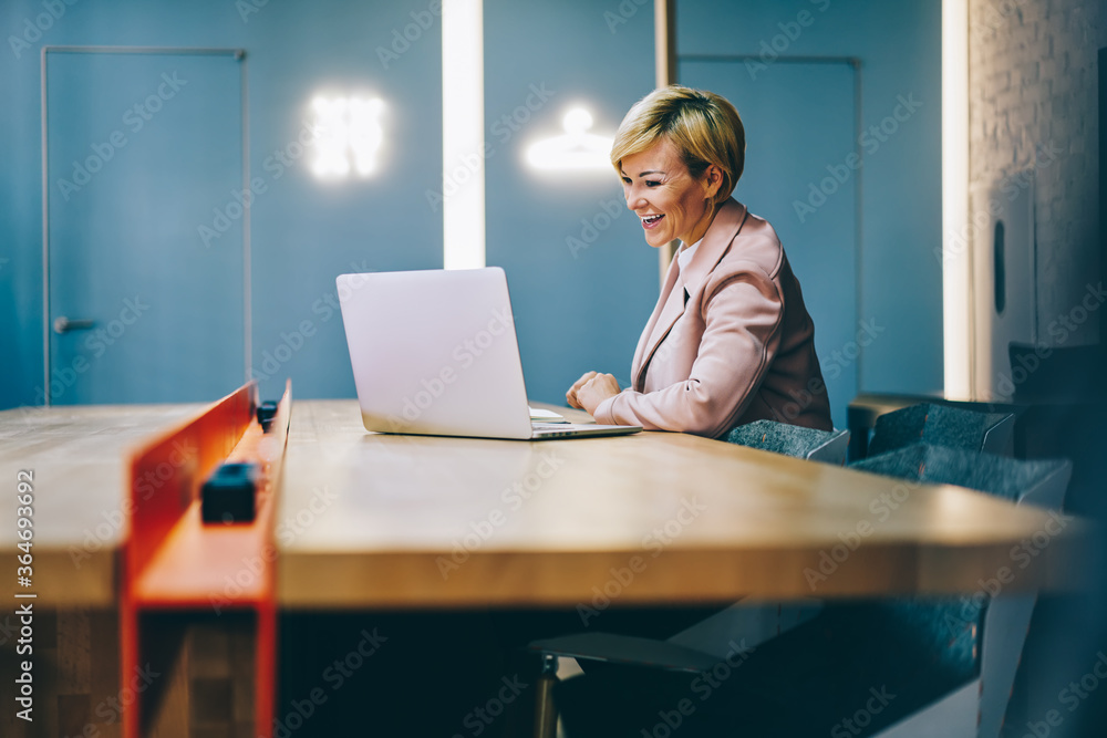 Positive mature coach laughing during watching business webinar online on modern laptop connected to wireless 4G internet.Cheerful female entrepreneur 50 years old reading funny news on netbook - obrazy, fototapety, plakaty 