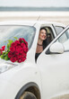 © Ann Stryzhekin - A girl with long black hairs developing in the wind with a bouquet of red roses on her white car in nature.