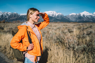 Wall Mural - Young female explorer observing breathtaking nature view enjoying hiking tour with backpack, cheerful girl wanderlust looking at scenery of mountains landscape spending leisure on discovering.