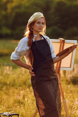  a girl artist with long red hair draws on an easel with a brush against the background of the sunset