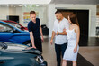© Andrii - A young couple chooses a new car at the dealership and consults with a representative of the dealership. Used cars for sale. Dream fulfillment