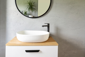 White washbasin with faucet on wooden countertop in minimalist modern bathroom. Scandinavian interior with stylish grey wall and round mirror. Copy space and nobody.