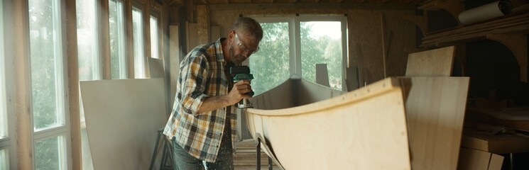 Wall Mural - Mid 50s Caucasian male building a canoe in his workshop, using milling tool. Boat making hobby, small business owner