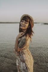 Wall Mural - Young female with tattoos wearing a dress and straw hat on blurred ocean background