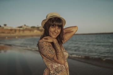 Wall Mural - Young female with tattoos wearing a dress and straw hat on blurred ocean background