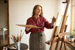 © shurkin_son - Beautiful elderly gray haired female artist working in her studio holding palette, applying acrylic paint on canvas using paintbrush, smiling. Inspiration, creative skills and art concept