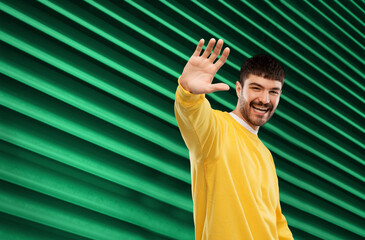 Wall Mural - gesture and people concept - smiling young man in yellow sweatshirt waving hand over emerald green background