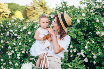 Young and beautiful mother holding baby girl toddler in white dress. Family in park spend time together, tender, love, hugs, play. Stylish summer clothing. 