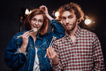 Young attractive couple - handsome man in checkered shirt and beautiful girl in denim jacket enjoys delicious sushi rolls in dark studio