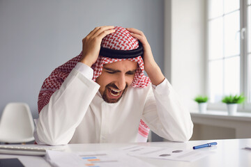 Wall Mural - Arab man holds his head with his hands while sitting at a table in the office.