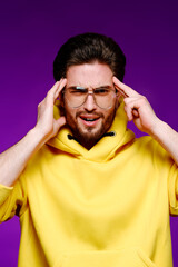 Wall Mural - A young man of 25-30 years in glasses and a yellow sweatshirt emotionally poses holding his head on a purple background.