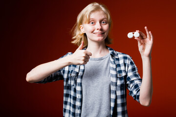 Blonde girl with bad eyesight keeps the container with lenses and shows the thumb up on red background