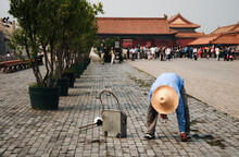 An Elderly Maintenance Man Picking Up The Trash From The Forbidden City Area, Wearing A Straw Hat And Blue Shirt, Tourists In The Background. 