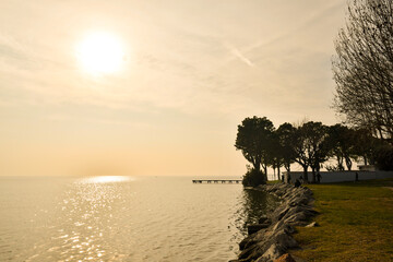 Wall Mural - Backlight view of the shore of Lake Garda with a pier and trees silhouettes at sunset, Bardolino, Verona province, Veneto, Italy