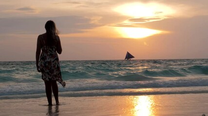 Wall Mural - Woman stands on beach barefoot wearing summer dress blowing in the wind and looking beautiful sunset. Lifestyle women travel on beach concept.