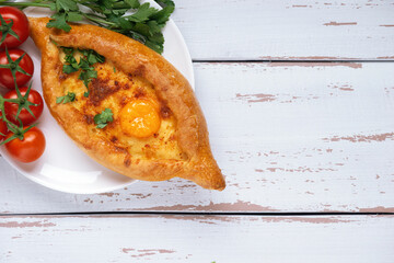 White plate with Adjarian Khachapuri – Georgian cheese bread decorated with tomatoes and parsley on white Provence style background. Baked open pie with cheese and egg yolk filling. Space for text.