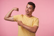 © timtimphoto - Cheerful young brown-eyed brunette male with short haircut smiling happily at camera while pointing with forefinger on his strong biceps, isolated over pink background