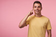 © timtimphoto - Happy young brown-eyed brunette male smiling cheerfully while listening to music and keeping finger on his earpiece, standing against pink background