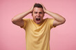 © timtimphoto - Stressed young brown-eyed short haired male clutching his head with raised hands and shouting angrily with wide mouth opened, standing over pink background