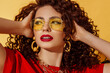 Close up summer fashion portrait of beautiful curly woman with fuchsia color lips makeup, wearing trendy yellow sunglasses, big earrings, colorful clothes, posing on yellow background