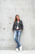 © perfectlab - Young cute woman is standing by the wall in a leather jacket, jeans and sneakers.