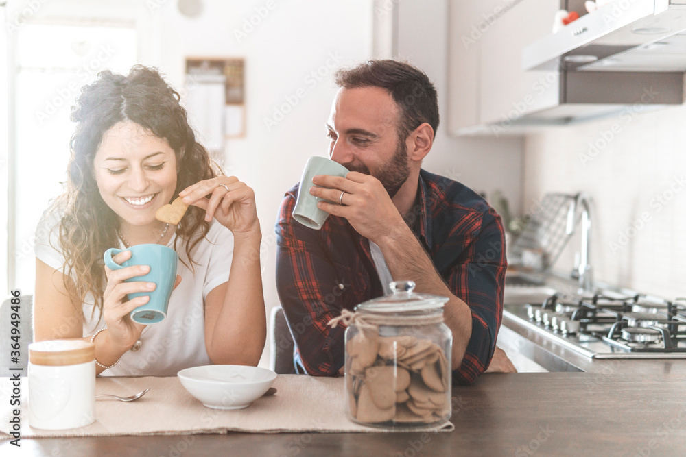 Engaged couple has breakfast together in their new home - young couple smiling while drinking and eating in the kitchen - warm filter on background - obrazy, fototapety, plakaty 