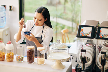 Wall Mural - Pretty smiling young Vietnamese woman decorating ice-cream in small bowl with sprinkles