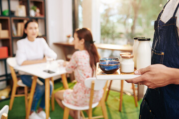 Wall Mural - Waiter bringing tray with delicious breakfast to pretty young women waiting at table