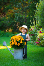 A Boy In A Green Jumpsuit And Hat Near A Watering Can With Yellow Daisies In The Garden During The Day