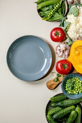 Wall Mural - Variety of fresh raw organic vegetables for cooking. Half of hokkaido pumpkin, young green pea, cauliflower, cucumbers and empty plate over beige background. Healthy eating concept. Flat lay, space