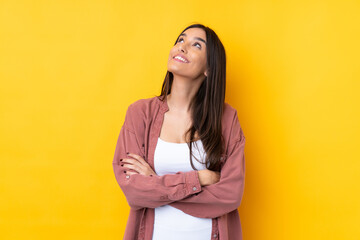 Young brunette woman over isolated yellow background looking up while smiling