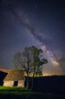 Old abandoned barn farm house with two trees next to it shot at night against a starry sky with milky way galactic core seen above