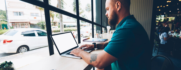 Serious bearded male blogger typing message on laptop computer with mock up screen chatting in social networks,pensve man checking mail via netbook spending time in cafe interior freelancing