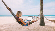 Female Freelancer Entrepreneur Lying On Hammock And Doing Remote Business Work At Modern Laptop Computer With Blank Screen Area For Web Page On Smartphone During Summer Vacation On Tropical Seashore