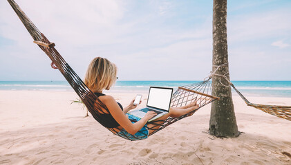 Female freelancer entrepreneur lying on hammock and doing remote business work at modern laptop computer with blank screen area for web page on smartphone during summer vacation on tropical seashore