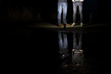 Two anonymus young men standing in front of a pool of water, during night time. Their reflection is hitting the water because of the hard flash light that is positioned in front of their feet.