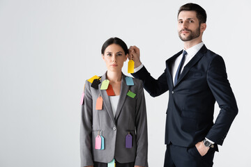 Wall Mural - bearded businessman standing with hand in pocket and putting label on beautiful businesswoman isolated on white, sexism concept