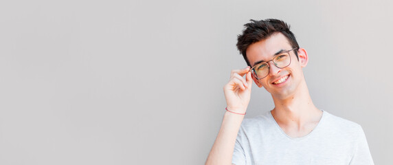 Wall Mural - A portrait of young handsome man in grey t-shirt isolated on gray background touching his glasses.