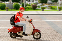 Young Guy Working As Deliveryman. Man Is Driving A Scooter