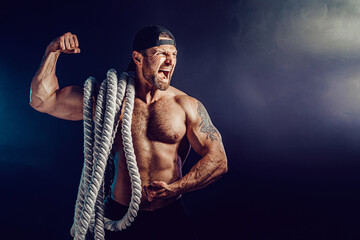 Aggressive bearded muscular sportsman holding battle rope on dark studio background with smoke. Strength and motivation.