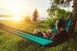© mykolastock - Modern male hipster holding smartphone, relaxing and enjoying listening to music from streaming service in hammock in park at sunset