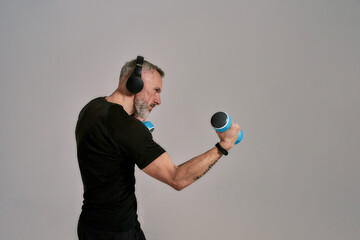 Wall Mural - Practice. Middle aged muscular man in black t shirt and headphones looking focused holding blue dumbbells, posing in studio over grey background