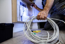 Close Up Shot Of Hand Of Aged Electrician, Repairman In Uniform Working, Fixing, Installing An Ethernet Cable In Fuse Box, Holding Flashlight And Cable