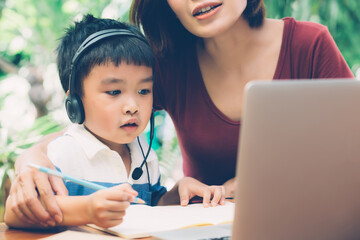 Wall Mural - Young asian mother and son using laptop computer for study and learning together at home, boy writing on notebook for homework and wearing headphone, teacher or mom support child, education concept.