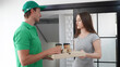 © FotoArtist - deliver man in green uniform handling bag of food, coffee cup give to female costumer in front of the house. Postman and express grocery delivery service during covid19.