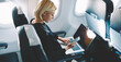 © BullRun - Businesswoman with short haircut sitting in airplane cabin and chatting online on smartphone while checking email on laptop computer with mock up area.Female traveler reading notification on cellular