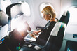 © BullRun - Blonde female tourist checking incoming notification on smartphone sitting on seat of airplane with netbook.Young businesswoman share media from telephone on laptop computer during plane flight