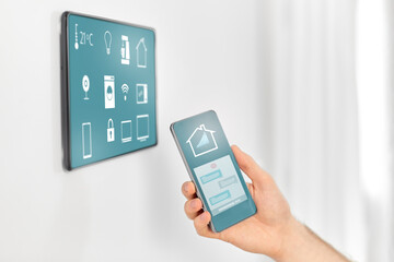 automation, internet of things and technology concept - close up of male hand with smartphone looking at tablet pc computer on wall with menu icons on screen at smart home