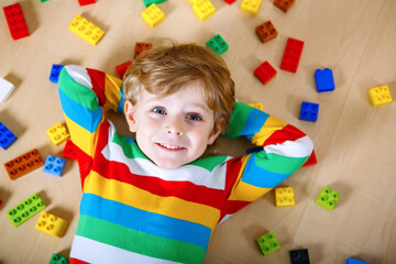 Little blond child playing with lots of colorful plastic blocks indoor. Kid boy wearing colorful shirt and having fun with building and creating